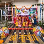 Group of children in colorful dresses posing on a decorated school stage with garlands umbrellas and a marigold rangoli